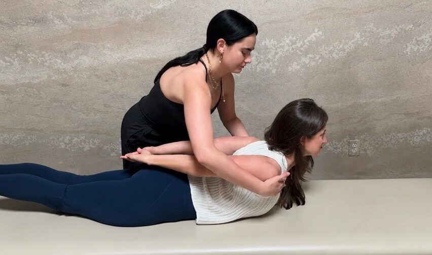 Emma kneeling beside a client, giving a hands-on assist during a prone back-extension exercise